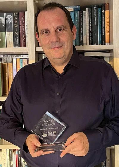Kostas holding his Friend of Darwin award in front of his bookshelf full of books